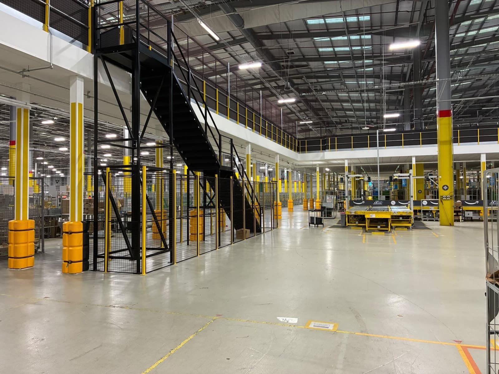 Warehouse floor beside the new mezzanine, with yellow and white columns under it and a black stair inside a mesh cage