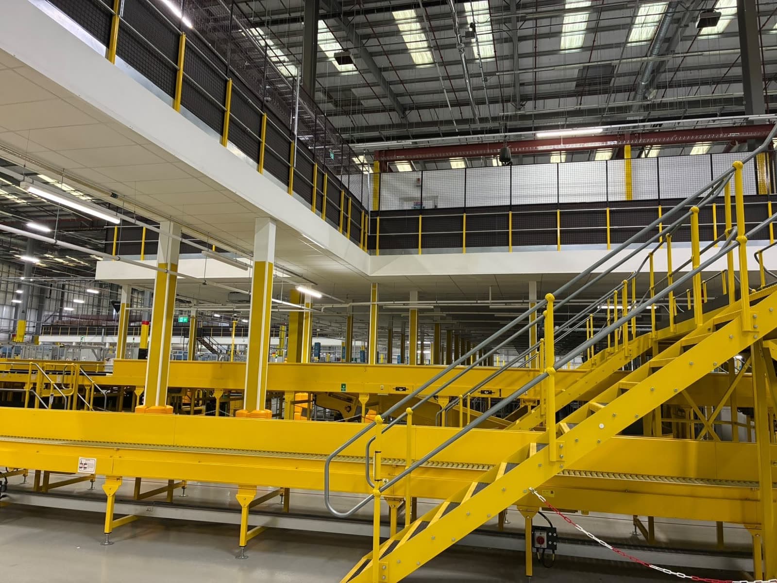 Yellow and white columns under the mezzanine, behind yellow conveyors and a yellow stair